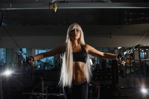 Unga muskulösa idrottskvinna utbildning med block simulator i gym, arbetar hårt för starka armmuskler. Flickan ser fram emot att skjuta händerna på sidorna. — Stockfoto