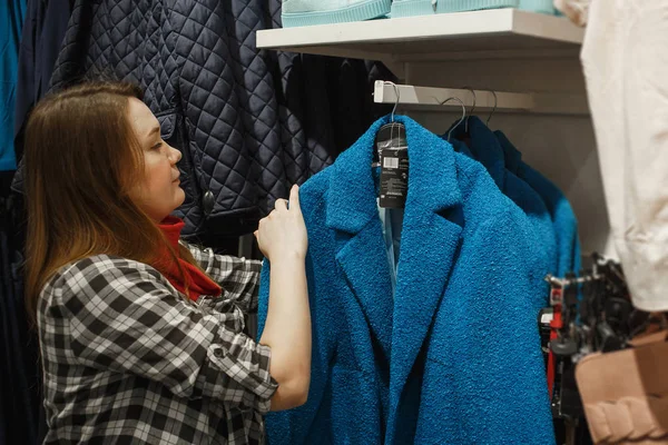 Shopping, moda, vendita, stile e concetto di persone - giovane donna che sceglie il cappotto blu nel negozio di abbigliamento — Foto Stock