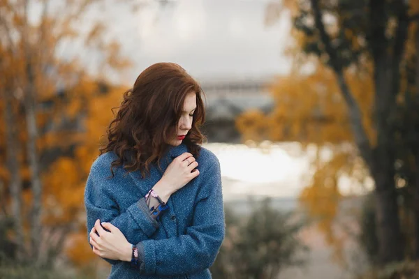 Trendy young woman in warm blue coat posing in the autumn park. She closing the coat by her hands — Stock Photo, Image