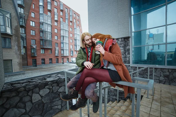 Молодая европейская пара веселится на зимнем городском фоне. Панки или хипстеры. Рыжая девушка одета в красный шарф, коричневое пальто, короткие шорты и колготки. Бледный человек одет в зеленое болоньезе — стоковое фото