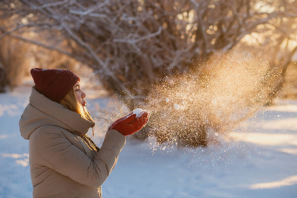 Winter Holiday, fun and people concept - young woman in red winter hat blowing snow from red mittens in sunny winter day
