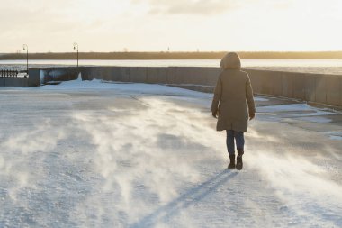 Kışın dondurucu bir nehir dolgu üzerinde gidiş bir kukuleta ile bir kış ceket kadın arkadan görünüm. Blizzard.