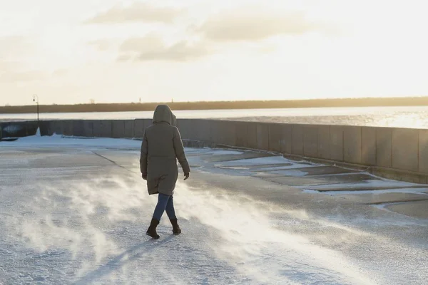 Back view woman in a winter jacket with a hood going on the embankment of a freezing river in winter. Blizzard. — Stock Photo, Image