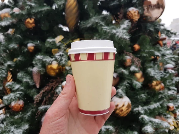 Tea or coffee in disposable cup in hand on Christmas Eve on the background of Christams tree with decorations.