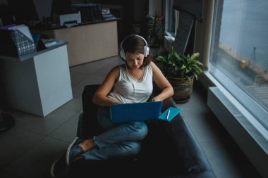 Kulaklık müzik dinlemek ve Office kanepede dizüstü bilgisayar üzerinde çalışan genç kadın. Rekreasyon, rahat iş kavramı