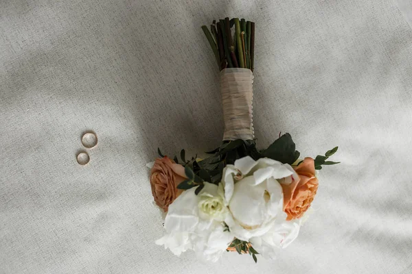Ramo de novia con rosas beige y cremosa y anillos de novia en la cama blanca — Foto de Stock
