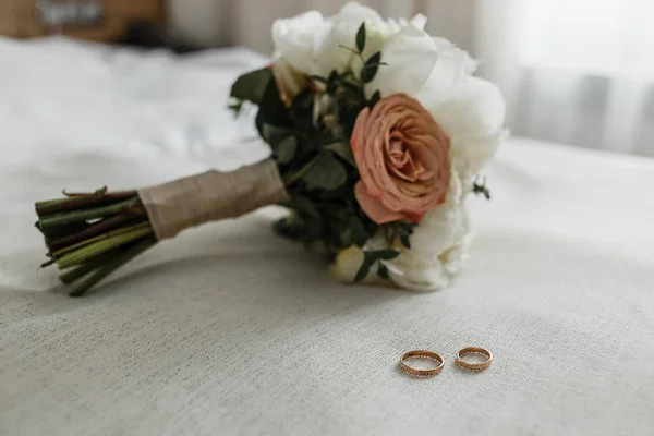 Ramo de novia con rosas beige y cremosa y anillos de novia en la cama blanca — Foto de Stock