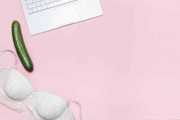 Erotic chat, virtual sex concept. White bra thrown on a modern laptop and big cucumber on a pink background. Minimalistic Flat lay, top view