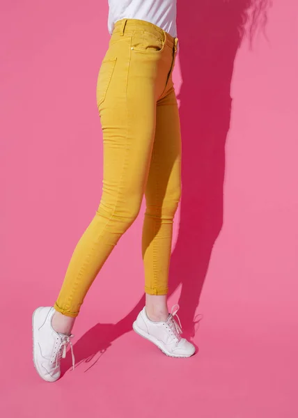 Женские ноги в модных желтых джинсах и белых кроссовках на розовом фоне — стоковое фото