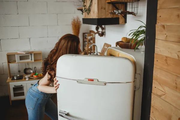 Smukłe dziewczyny w dżinsy otwarcie retro drzwi lodówki i patrząc w lodówce — Zdjęcie stockowe