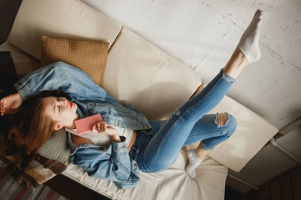 Cute young woman in jeans clothes lying in sofa with pink diary — Stock Photo, Image