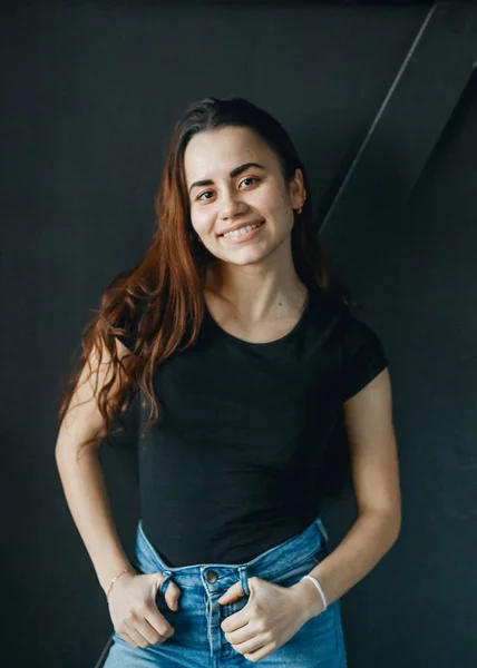 Mujer joven y feliz mirando a la cámara en la pared negra. Ella se vistió con camiseta negra y jeans — Foto de Stock