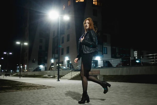 Таинственная рыжая женщина в элегантной кожаной куртке и на высоких каблуках ходит по городской улице, нуар атмосфера, интригующая концепция — стоковое фото