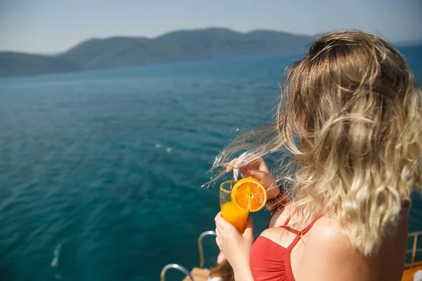 Счастливая грудастая женщина на яхте. Девушка в коралловом бикини со свежим апельсиновым напитком. Прохладный напиток и свежее аквамариновое море. Жить счастливой жизнью — стоковое фото