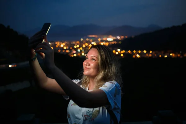 Jeune femme prenant selfie au sommet de la colline observant la vue nocturne sur la ville. Bonne lumière de l'écran — Photo