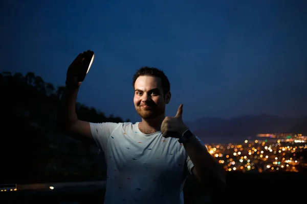 Ung man tar Selfie på toppen av kullen Observera natten stadsutsikt. Bra ljus från skärmen. Tummen upp gest — Stockfoto