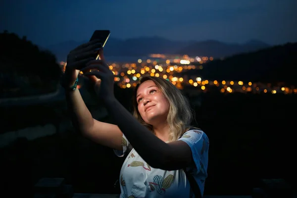 Jeune femme prenant selfie au sommet de la colline observant la vue nocturne sur la ville. Bonne lumière de l'écran — Photo