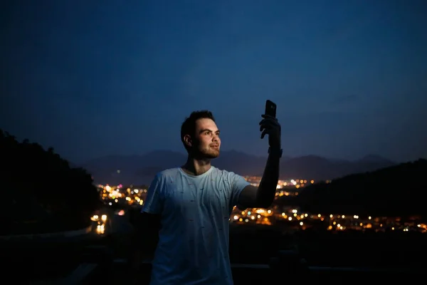 Молодой человек делает селфи на вершине холма, наблюдая ночной вид на город . — стоковое фото