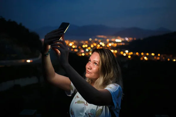 Jeune femme prenant selfie au sommet de la colline observant la vue nocturne sur la ville. Bonne lumière de l'écran — Photo