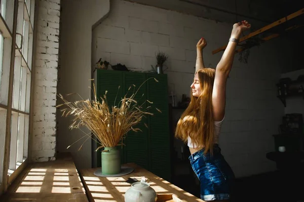 Söt ingefära kvinnlig modell i fritidskläder njuter av morgonen. Trevlig ingefära Kvinna lyfta händerna i solljus från fönster — Stockfoto