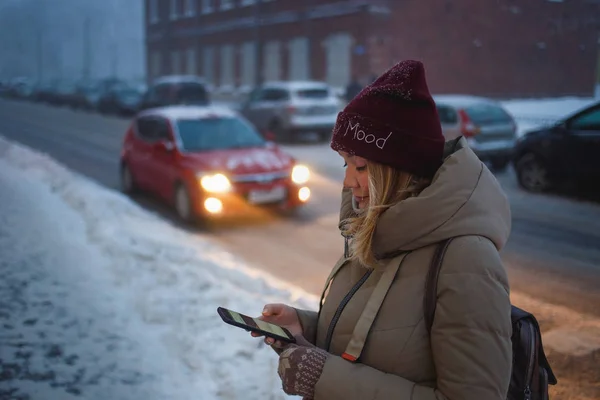 Jonge mooie vrouw met behulp van haar mobiele telefoon in de schemering in de stad. Lachend meisje belt taxi van mobiele telefoon in stedelijke omgeving. Vrouw surfen op het net met smartphone op een winterdag in de straat — Stockfoto