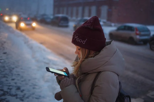 Ung vacker kvinna som använder sin mobiltelefon i skymningen i staden. Leende flicka ringer taxi från mobil i stadsmiljö. Kvinna surfa på nätet med smartphone på en vinterdag på gatan — Stockfoto