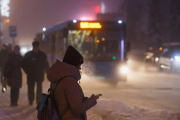 Jonge vrouw het controleren van de smartphone tijdens het wachten op de bus te komen. Froty winter. Schemer tijd. — Stockfoto