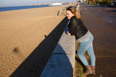 Sonbaharda dışarı bakan mutlu, kıvrımlı genç bir kadın. Kız güneşli havanın tadını çıkarıyor. Kot pantolon ve siyah deri ceket giyiyor.