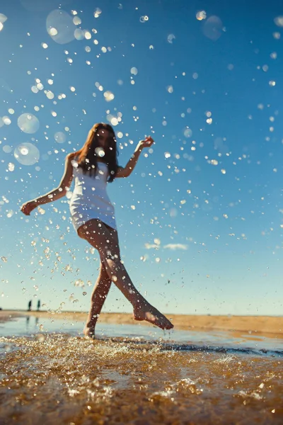 Молодая стройная девушка в белом платье бежит по воде с брызгами на солнце. Концепция летнего отдыха на пляже, хорошая погода, счастье — стоковое фото