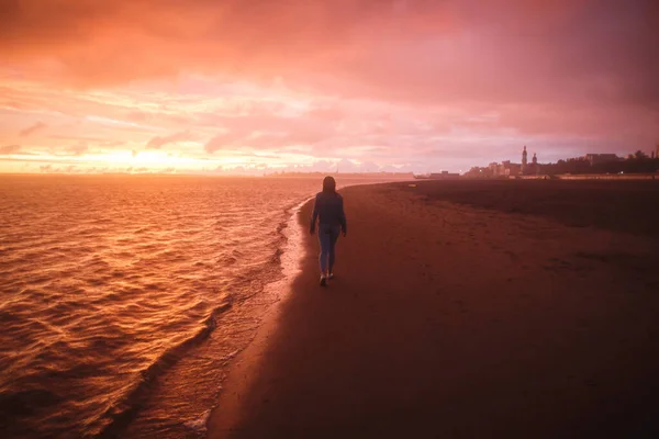 Una vista desde detrás de una chica camina sola en la playa de la ciudad y mira el colorido atardecer después de la lluvia. Naranja, morado carmesí — Foto de Stock