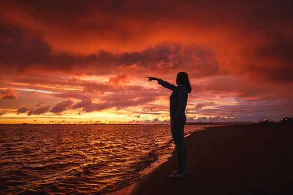 Woman walks alone on the beach and looks at the colorful sunset after the rain. Legs in the white shoes. Red orange, crimson purple colours