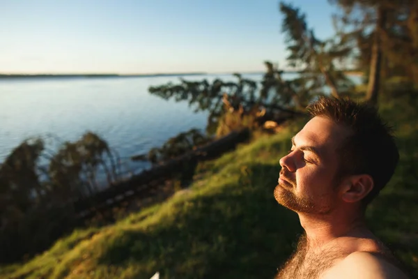 Красивий бородатий білий чоловік із закритими очима, що стоять на скелі на красивому заході сонця. На задньому плані річка і тайга ліс — стокове фото