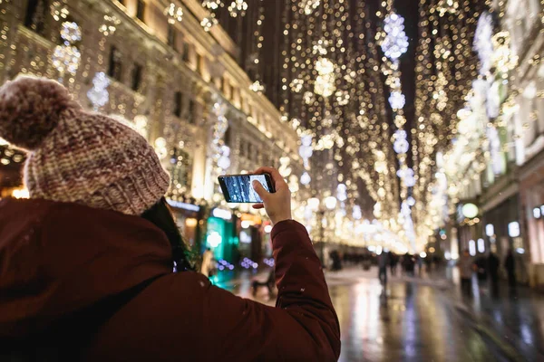 Outdoor night portrait of young fashionable woman in the winter hat with pompom taking photo by phone rear view. Magic snowfall effect. Night street illumination in Moscow