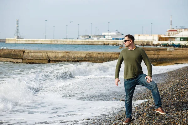 Красивий чоловік в окулярах і зелений светр спостерігає на скелястому пляжі в холодну пору року. Сезонні, люди та концепція способу життя — стокове фото