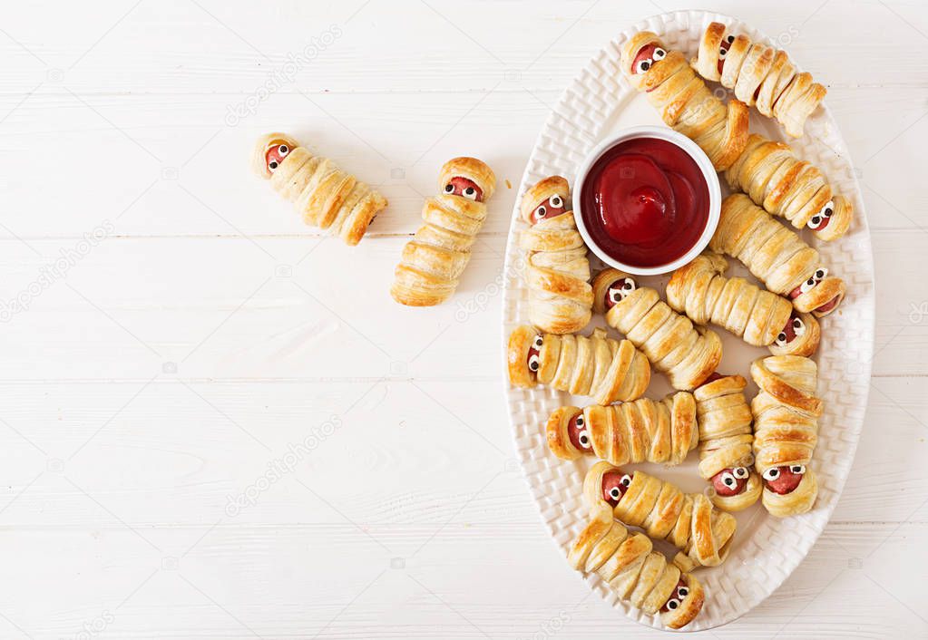 Scary sausage mummies in dough with funny eyes on table. Funny decoration. Halloween food. Top view. Flat lay