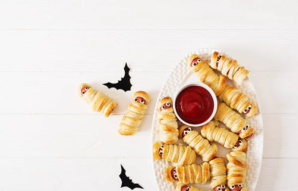 Scary sausage mummies in dough with funny eyes on table. Funny decoration. Halloween food. Top view. Flat lay