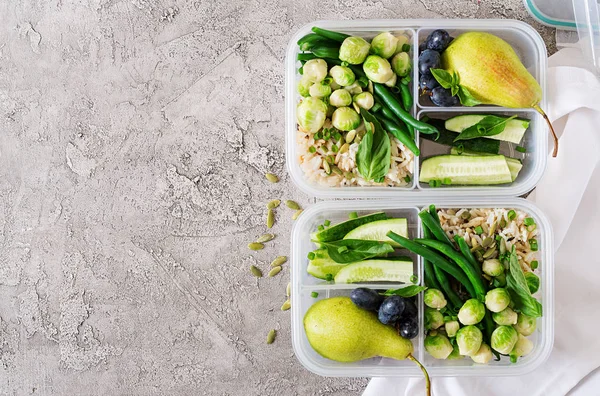Vegan green meal prep containers with rice, green beans, brussel sprouts, cucumber and fruits on stone grey background