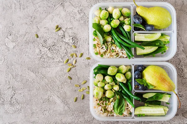Vegan green meal prep containers with rice, green beans, brussel sprouts, cucumber and fruits on stone grey background
