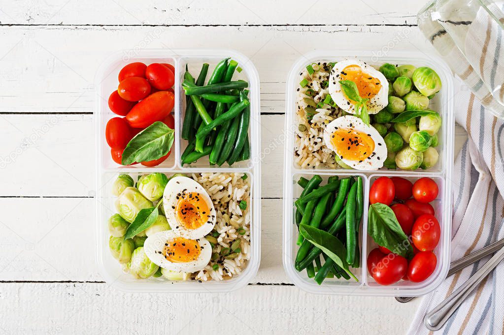 Vegetarian meal prep containers with eggs, brussel sprouts, green beans and tomatoes on white wooden background