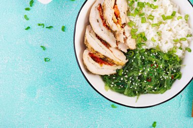 Beyaz bir kapta sağlıklı salata, yemek çubukları. Tavuk rulo, pirinç, chuka ve yeşil soğan. Mavi arka plan. Asya mutfağı. Üstte görüntü