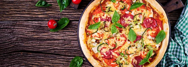 Italian pizza with chicken, salami, zucchini, tomatoes and herbs on vintage wooden background. Top view. Banner. Italian cuisine