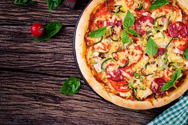 Italian pizza with chicken, salami, zucchini, tomatoes and herbs on vintage wooden background. Top view. Italian cuisine