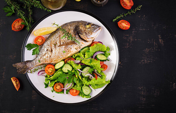 Baked fish dorado with lemon and fresh salad in white plate on dark rustic background. Top view. Healthy dinner with fish concept. Dieting and clean eating