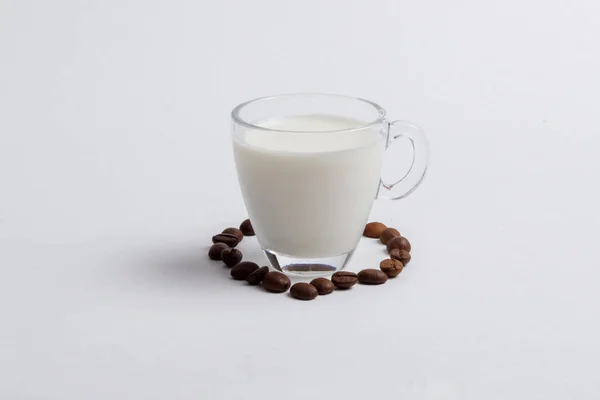 A glass mug full of milk is standing among the coffee grains in the form of a heart on a white background