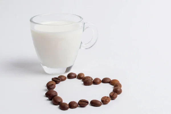 A glass mug full of milk is standing next to coffee beans in the form of a heart on a white background