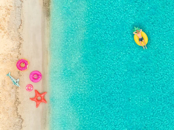 Aerial view of man floating on inflatable pineapple mattress by sandy beach and inflatable rings.