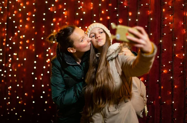 Mother kissing cheek of daughter during selfie at Christmas market.