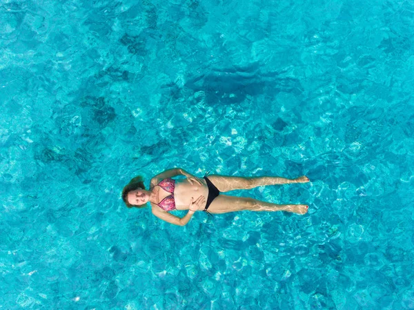 Aerial view of pregnant woman floating on her back in transparent sea holding her tummy.