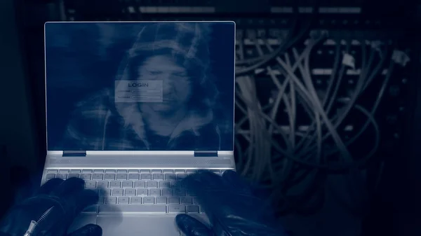 Hacker with laptop on a dark background of structured cabling. Close-up of male hands in black gloves on keyboard, motion blur. Mysterious reflection of a spy\'s head in hood on a digital display.