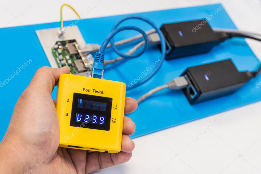 Measurement of PoE power voltage by data cable tester in technician hand. Human palm holding yellow diagnostic digital gauge. Two switched supplies, electric wires and surge protector on blue plastic.